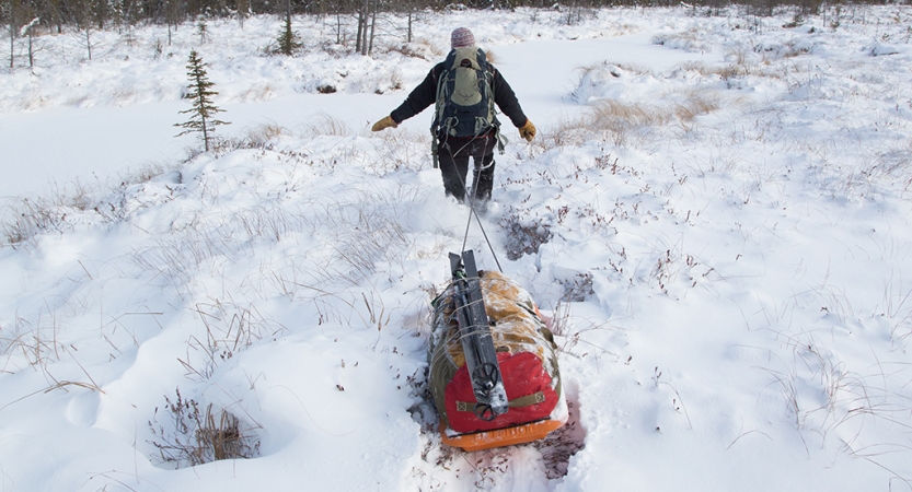 A person wearing a backpack pulls a small sled behind them through deep snow